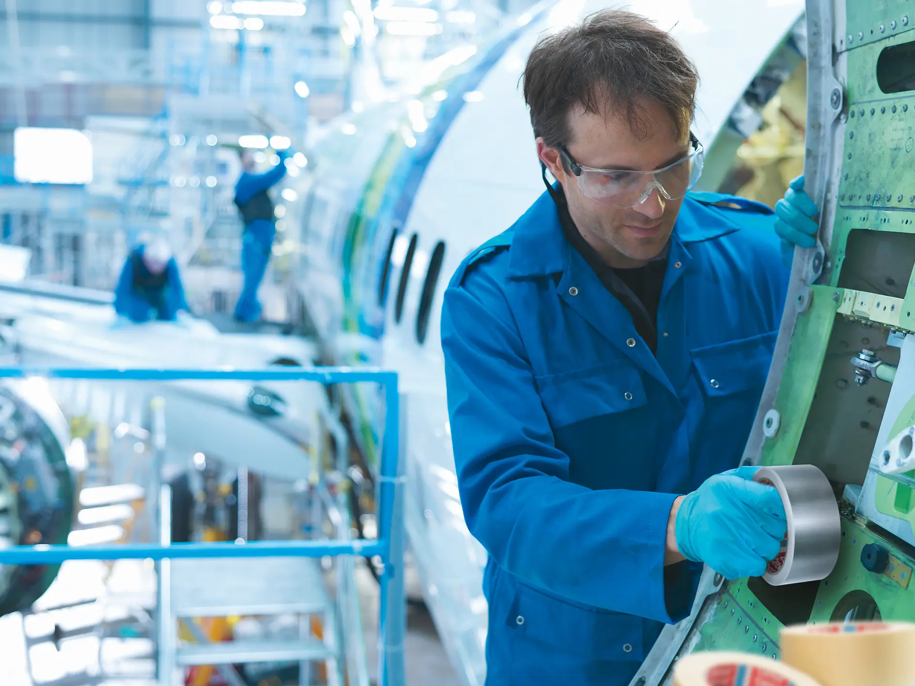 tesa tape being applied by a technician in blue coveralls inside an aircraft maintenance hangar with airplane parts visible in the background