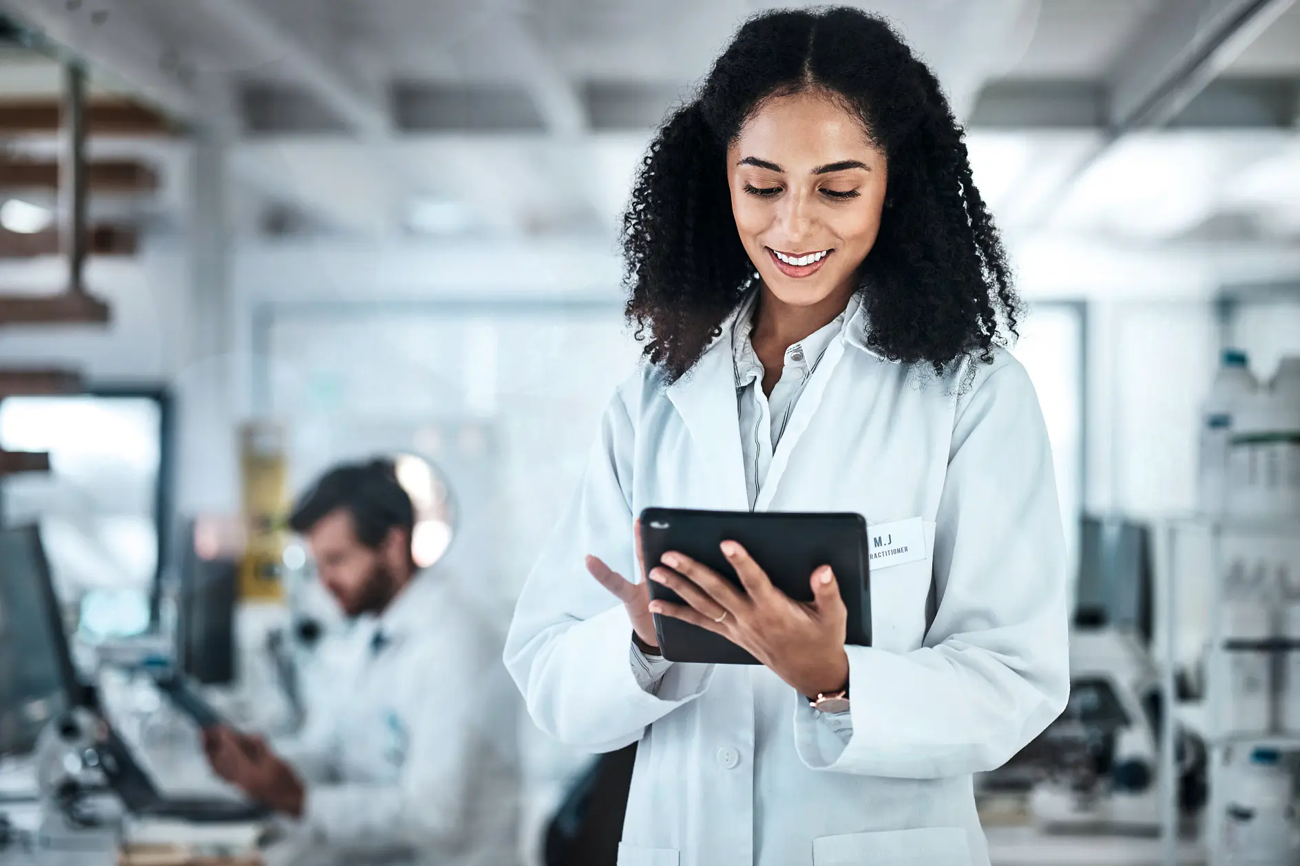 scientist in white lab coat holding tablet in modern laboratory with colleague working on equipment in background (This text has been generated by AI)