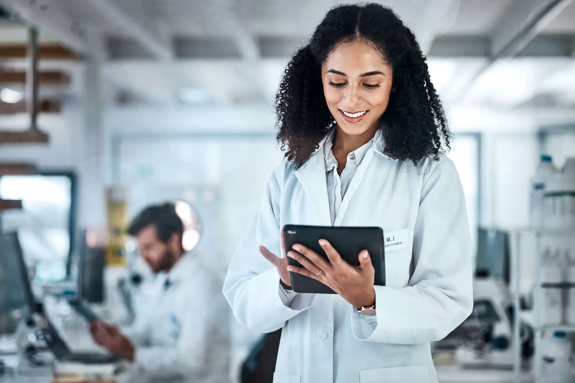 scientist in white lab coat using tablet in modern laboratory with colleague working in background