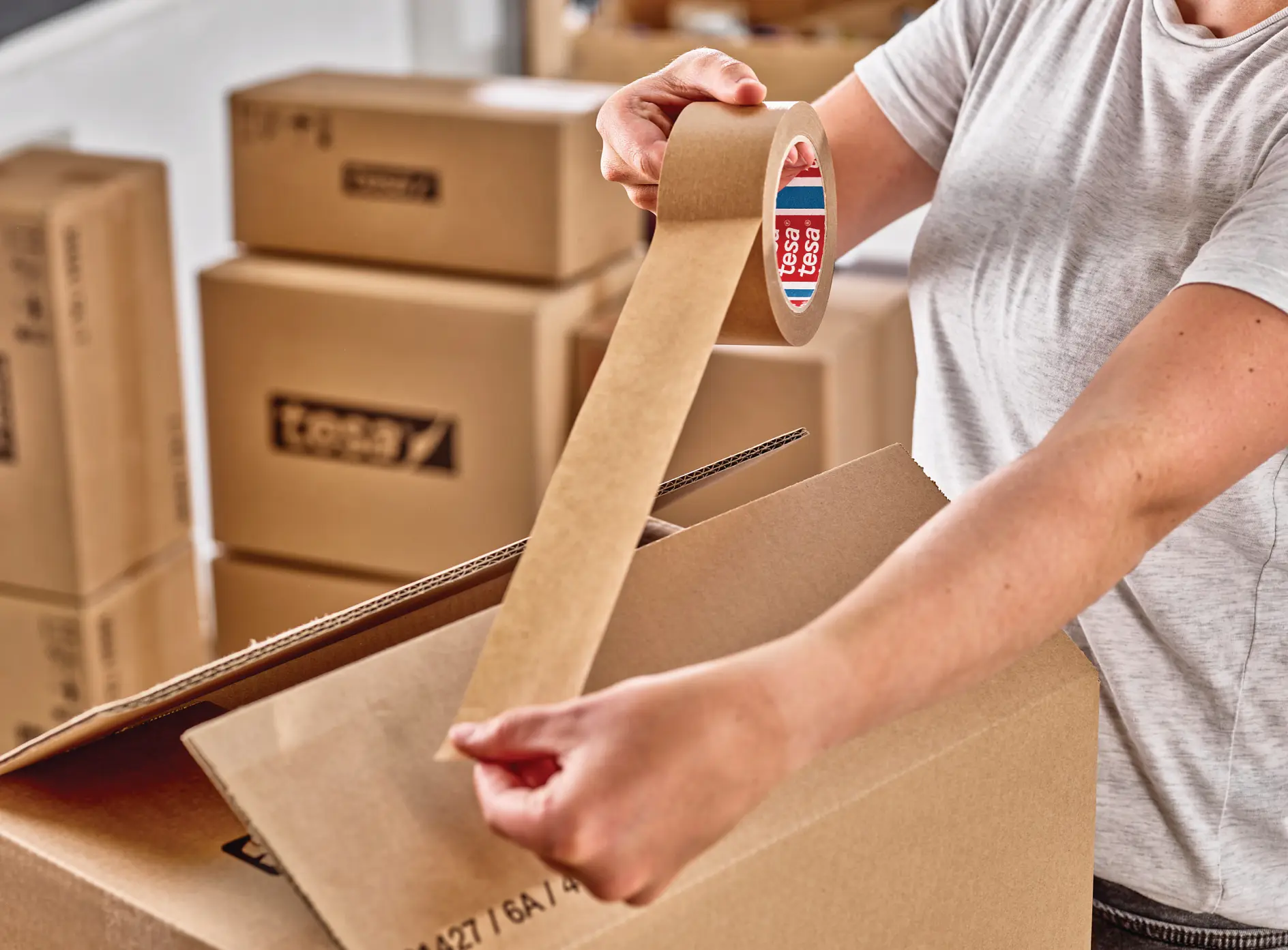 person sealing a cardboard box with brown packing tape in a room filled with stacked moving boxes