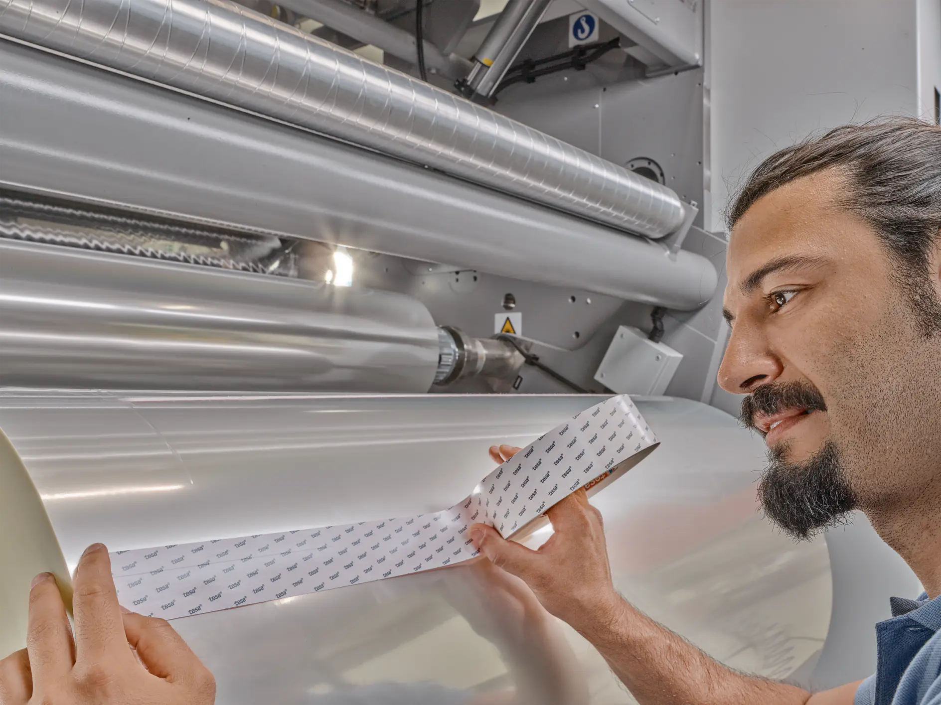 man inspecting a roll of transparent adhesive film in an industrial setting with large metal rollers and machinery in the background (This text has been generated by AI)