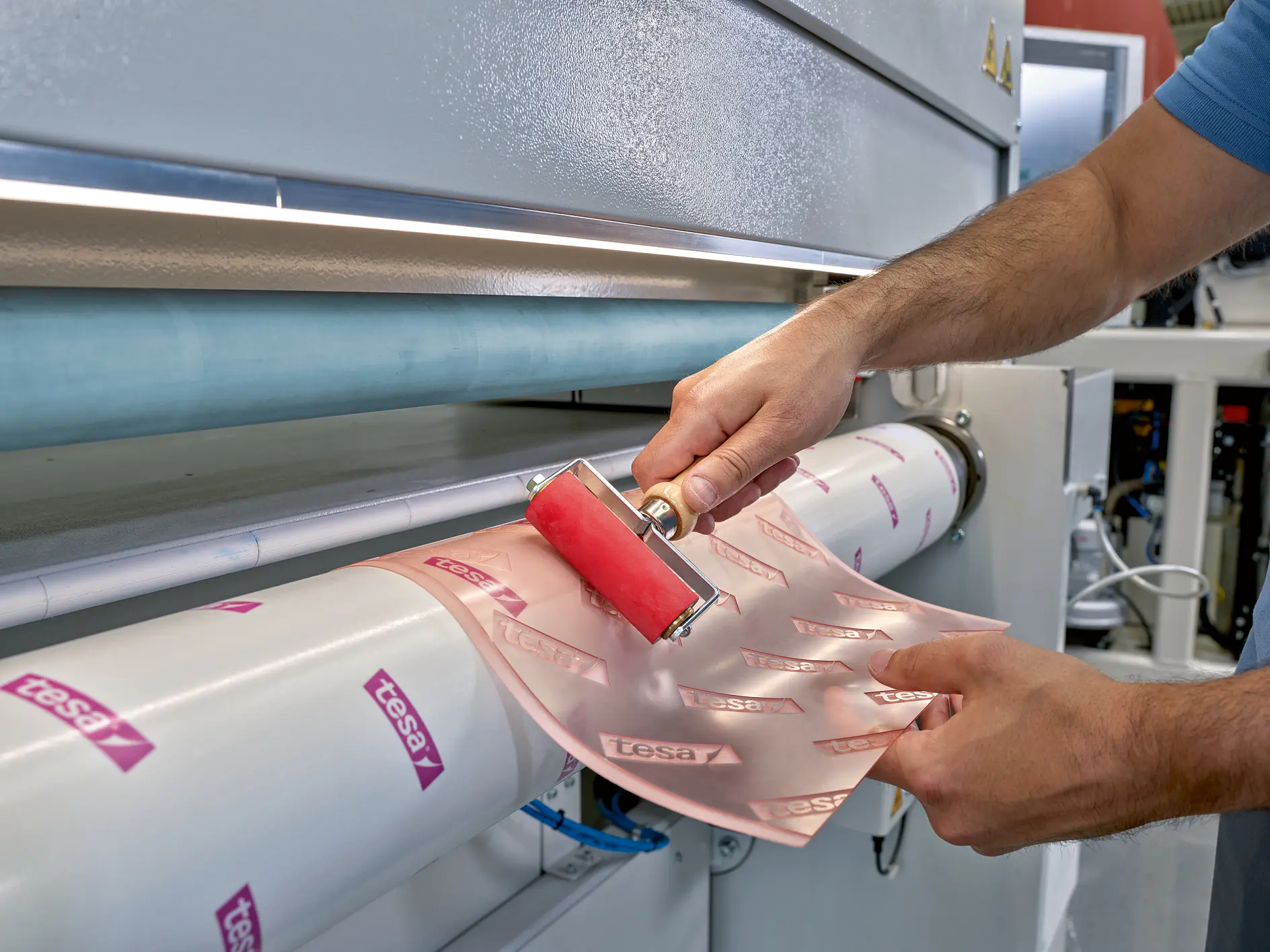 hands applying tesa branded adhesive film onto a large roll in an industrial printing or laminating machine indoors (This text has been generated by AI)