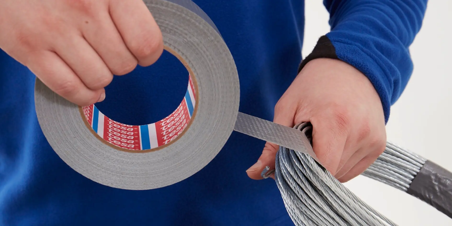 hands wrapping silver adhesive tesa tape around a bundle of metal wires against a white background