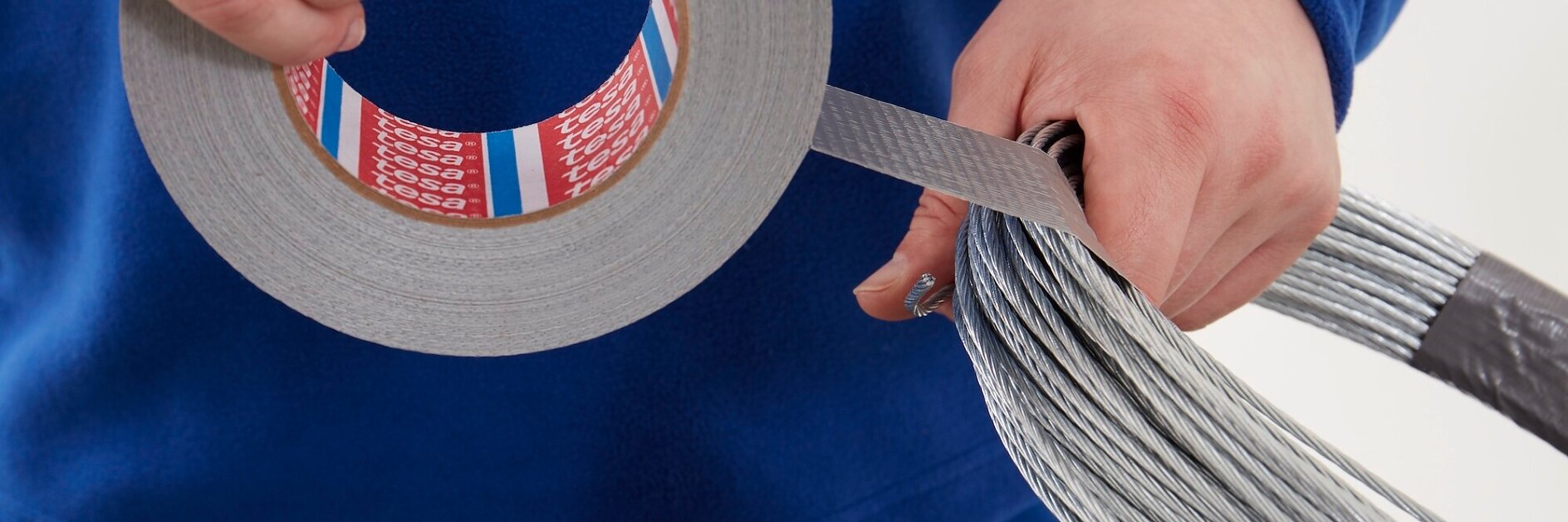 hands wrapping silver adhesive tesa tape around a bundle of metal wires against a white background