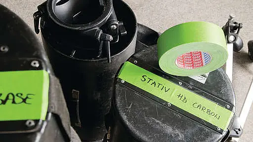 黑色圆柱形盒子特写，盒子上贴有“Stativ HD Carbon”标签并用绿色tesa胶带固定，顶部放有一卷同款绿色tesa胶带。盒子置于灰色表面上，显示出设备安装场景。