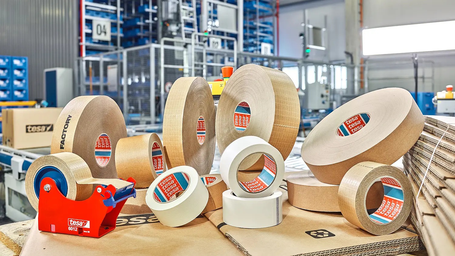 various rolls of tesa adhesive tape and tape dispensers arranged on cardboard in a warehouse setting with industrial shelving in the background