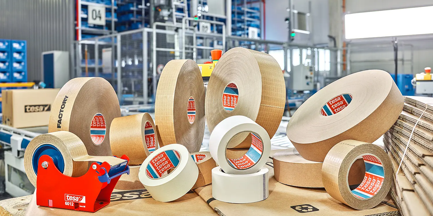 various rolls of tesa adhesive tape and tape dispensers arranged on cardboard in a warehouse setting with industrial shelving in the background