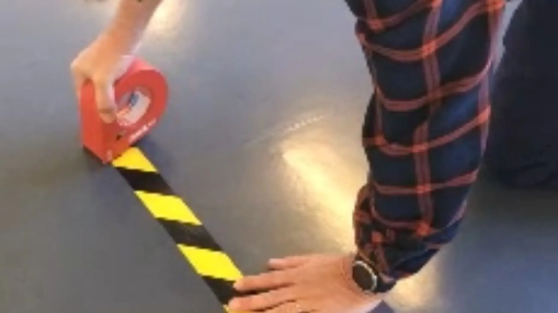 一位穿格子衬衫的人正在使用红色的tesa胶带，将一条黑黄相间的警示tesa胶带贴在地板上。他们正跪着，并在左手腕上戴着手表。