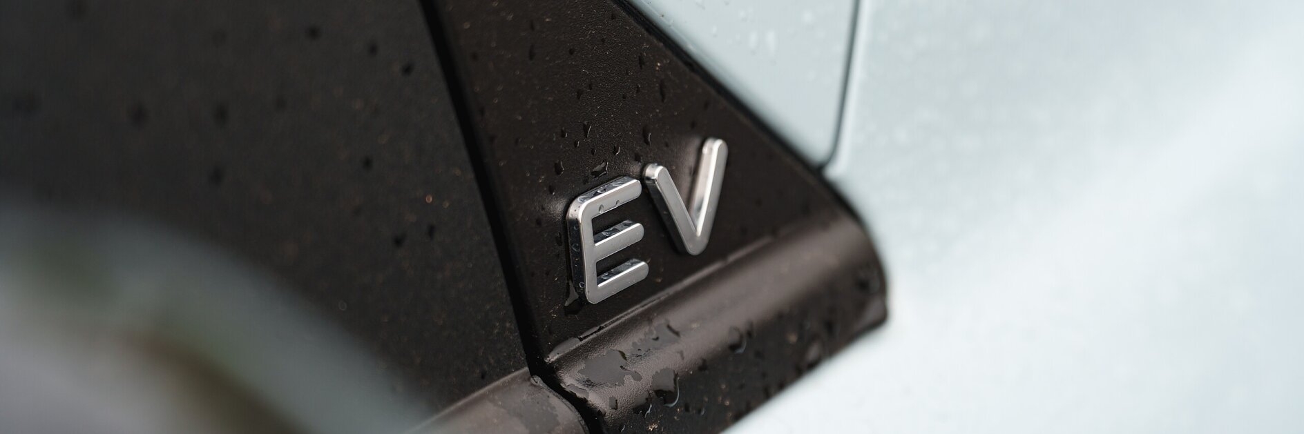 Close-up of a car with silver EV lettering on a black panel. The light blue body and the surface are covered in water droplets, indicating recent rain. tesa tape has been used in the assembly or detailing, though only a portion of the vehicle is visible.